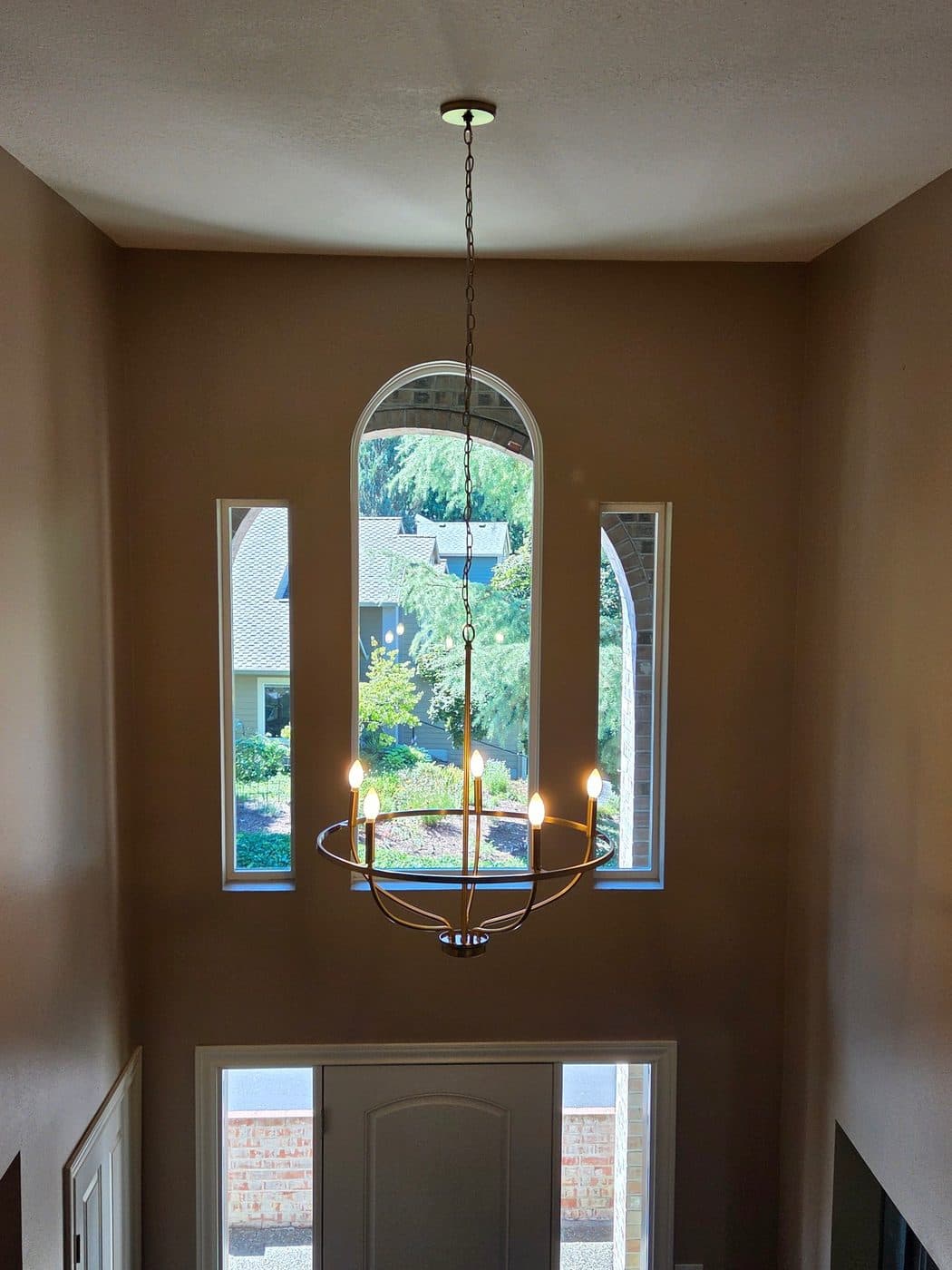 A brass ring chandelier hanging on a long chain in a double-height entry hall above an arched window